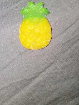 Gelégodis i form av ananas - Säljer ett gelégodis format som en ananas, med gul kropp och gröna blad. Godiset är täckt av socker och har en mjuk konsistens. Perfekt för dig som gillar fruktiga smaker och roliga former på godis.