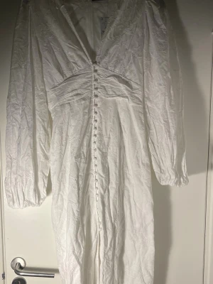 Vit långklänning med broderade blommor - Skriv privat för bild på! Ny klänning med prislapp på. Nypris 700. Perfekt till studenenten, konfirmation eller midsommar tex. Elegant vit långklänning med v-ringning, långa ärmar och dekorativa knappar framtill. Klänningen har ett subtilt blommigt broderat mönster.