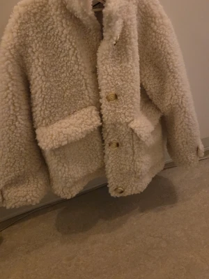 Offwhite teddyjacka från indiska - Supermysig teddyjacka från indiska i offwhite med stora fickor framtill och knappar i guldton. Jackan har en boxy passform och fluffigt material som ger en trendig look. Perfekt för att hålla sig varm och snygg under kyliga dagar. Aldrig använd.  nypris 899