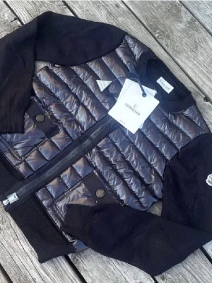 Svart pufferjacka från Moncler - Snygg svart pufferjacka från Moncler med glansig quiltad front och stickade ärmar. Jackan har två fickor med knappstängning, ribbade muddar och dragkedja framtill. Klassisk Moncler-logga på ärmen och en modern, sportig look.