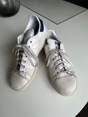 Adidas Stan Smith vita sneakers - Klassiska vita Adidas Stan Smith sneakers med mörkblå detalj på hälen och perforerade ränder på sidorna. Skorna har lågt skaft, vita snören och ovandel i läder. Perfekta för en clean och tidlös stil.