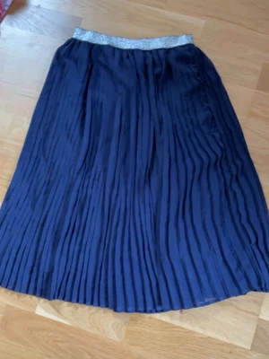 Mörkblå plisserad långkjol med glitter - Snygg mörkblå plisserad långkjol med bredt glittrigt silverresår i midjan. Kjolen har ett elegant fall och passar perfekt till både vardagliga och mer uppklädda tillfällen. Materialet känns lätt och följsamt.