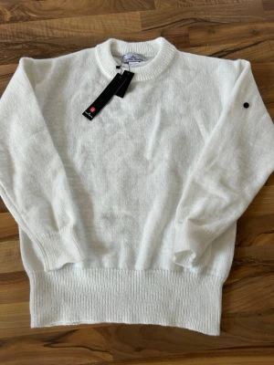 Vit stickad tröja från Stone Island - Säljer en stilren vit stickad tröja från Stone Island med rund halsringning och ribbade muddar. Tröjan har en diskret svart logodetalj på ärmen och är tillverkad i mjukt material som känns lyxigt. Perfekt för dig som gillar minimalistisk och clean stil. Helt oanvänd och tappade märket direkt men det finns att köpa för 50kr