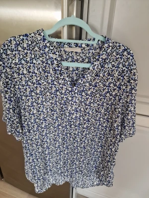 Blommig blus från Free Quent  - Snygg blus från Free Quent med korta ärmar och v-ringning. Mönstret är småblommigt i blått, svart och vitt på en ljus botten. Luftigt material som känns lätt och bekvämt. Perfekt till jeans eller kjol för en fräsch look. Använd två gånger bara.  I mycket gott skick.