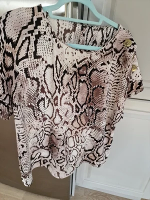 Topp från River Island  - Säljer en snygg topp med ormmönster i beige, brun och svart. Toppen har korta ärmar och tre dekorativa knappar på axeln som cool detalj. Använd 3 gånger bara. I mycket gott skick.