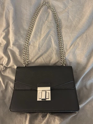 Svart axelväska med kedjerem - Snygg svart axelväska i slätt konstläder med silverfärgad kedjerem och stilrent metallspänne framtill. Väskan har en rektangulär form och minimalistisk design som passar till många olika looks. Perfekt för dig som gillar trendiga accessoarer med en clean vibe. Väskan är nästan aldrig andvänd, pris kan diskuteras.