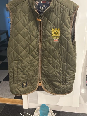 Olivgrön quiltad väst från Morris - Stilren olivgrön quiltad väst från Morris med guldfärgat broderat emblem och brittisk flagga på bröstet. Västen har bruna detaljer, dragkedja och träknapp upptill samt två fickor framtill. Perfekt för lager-på-lager och snygg till streetwear.