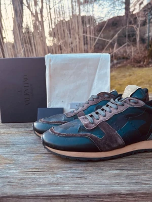 Valentino rockrunner - Säljer ett par snygga Valentino sneakers med camo-mönster i mörkgrönt och svart, detaljer i grå mocka och bruna partier. Skorna har coola nitar på hälen och en beige sula med svart yttersula. Perfekt för dig som vill sticka ut med exklusiva skor.