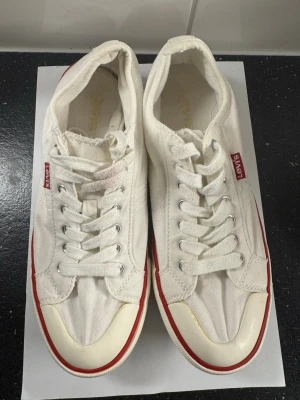 Vita Levi's canvas sneakers - Snygga vita sneakers från Levi's i klassisk canvas med vita snören och metallöljetter. Skorna har en låg profil och en röd rand längs sulan samt den ikoniska Levi's-taggen på sidan. Perfekta för en clean och avslappnad stil.