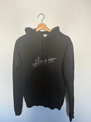 Svart hoodie från Saint Laurent - Säljer en svart hoodie från Saint Laurent med coolt tryck framtill där det står 'Saint Laurent' i graffiti-stil och en stjärna. Hoodien har huva med dragsko och en klassisk känguruficka. Perfekt för dig som gillar streetwear och exklusiva märken. Tröjan är köpt på miinto och har även kvitto på de, hör av er för fler bilder eller vid minsta funderingar 