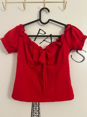 Röd omlott blus med puffärm från NLY Trend  - Röd blus med korta puffärmar och omlott-detalj framtill med knyt i storlek XS från NLY Trend. Toppen har en söt volangkant längs halsringningen och är figurnära i modellen.
