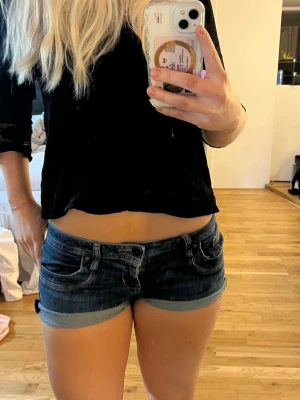 Lågmidjade jeansshorts  - Säljer ett par mörkblå jeansshorts med upprullad kant och klassisk femficksdesign. Shortsen har låg midja och är tillverkade i bomull med en liten mängd stretch för extra komfort. Perfekta för varma sommardagar! Storlek S/M/L för de är stretchiga så passar nog alla. Själv har jag M