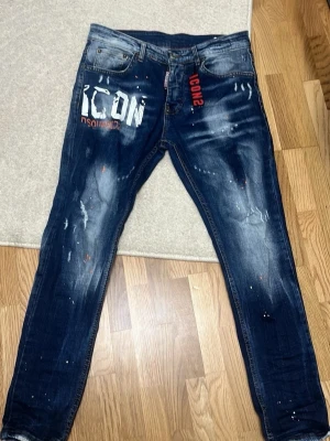 Mörkblå Dsquared2 ICON jeans - Säljer ett par mörkblå  jeans från Dsquared2 passar till sommaren med stor vit och röd ICON-logga framtill. Jeansen har slitningar, färgstänk och en tvättad look för en riktigt trendig streetstyle. Klassisk femficksmodell och råa benslut. 