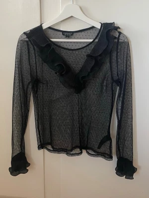 Svart meshblus med volang från Topshop - Säljer en transparent svart meshblus från Topshop i storlek 36. Blusen har prickigt mönster, långa ärmar och volangdetaljer längs ringningen och ärmsluten. Perfekt för dig som gillar trendiga och unika toppar!