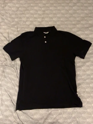 Svart pikétröja från EastWest - Klassisk svart pikétröja från EastWest med krage och tre vita knappar framtill. Tröjan har korta ärmar och är tillverkad i ett mjukt bomullsmaterial som känns skönt mot huden. Perfekt till jeans eller shorts för en clean look.