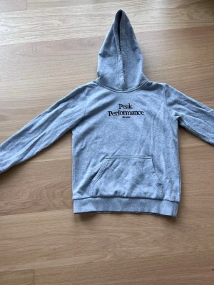 Grå original hoodie från Peak Performance - Snygg gråoriginal hoodie från Peak Performance med svart broderad logga och text 'Est. 1986' på bröstet. Klassisk känguruficka framtill och ribbade muddar. Mjuk bomullsblandning och skön passform, perfekt för chill dagar.