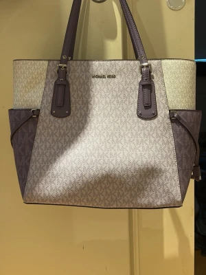 Michael Kors lila handväska - Stilren handväska från Michael Kors i lila med klassiskt MK-mönster över hela väskan. Väskan har två långa handtag i lila läder och detaljer i samma färg. Perfekt för dig som gillar lyxig och tidlös design. Tillverkad i tåligt konstläder.