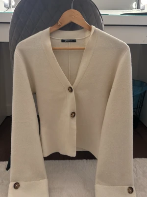 Krämvit kofta från Gina Tricot - Stilren krämvit kofta från Gina Tricot med v-ringning och två stora bruna knappar framtill. Koftan har långa ärmar med breda muddar och en mjuk, lite tjockare stickad känsla. Perfekt att slänga över en topp för en chill look.