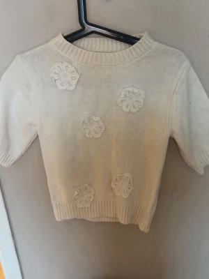 Vit stickad tröja med blommor - Söt vit stickad tröja med korta ärmar och dekorativa virkade blommor framtill. Tröjan har ribbad halsringning, ärmslut och nederkant. Perfekt för dig som gillar en gullig och stilren look.