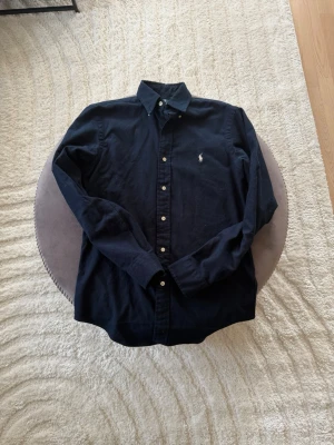 Mörkblå skjorta från Ralph Lauren - Klassisk mörkblå skjorta från Ralph Lauren med button-down krage och vita knappar. Skjortan har lång ärm och den ikoniska broderade loggan på bröstet. Perfekt till jeans eller chinos för en clean och stilren look.