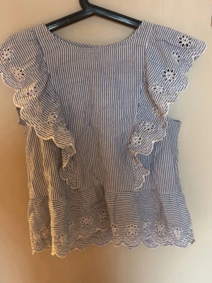 Blårandig blus med volang och brodyr - Söt blus med blåvita ränder, volangdetaljer och broderade blommor längs ärmar och nederkant. Blusen har rund halsringning och är tillverkad i ett lätt, luftigt tyg som passar perfekt till sommaren.