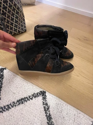 Isabel marant sneakers - Unika sneakers med kilklack i svart och brunt mocka-liknande material. Skorna har breda snören, detaljer i metall och en beige gummisula. Snygg design med högt skaft som ger extra edge till din outfit. Isabel maran Bobby