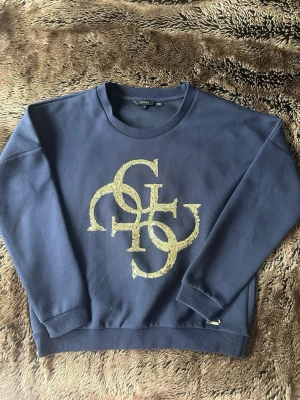 Mörkblå sweatshirt från Guess - Mörkblå sweatshirt från Guess med stort glittrigt guldtryck på bröstet. Tröjan har rund halsringning, ribbade muddar vid ärmslut och nederkant samt en avslappnad passform. 