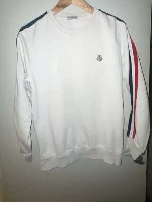Vit sweatshirt från Moncler - Vit sweatshirt från Moncler med rund hals och långa ärmar. Tröjan har röd och marinblå rand längs ärmarna samt Moncler-logga på bröstet. Tillverkad i mjuk bomull för en bekväm känsla. Perfekt för en sportig och stilren look.