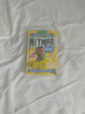 Neymar äger - Fotbollsstjärnor - En färgglad bok med titeln 'Neymar äger' från serien Fotbollsstjärnor, skriven av Simon Mugford och Dan Green. Omslaget är gult med illustrationer och fotbollstema. Boken är inbunden och innehåller svartvita illustrationer och fakta om fotbollsspelaren Neymar.