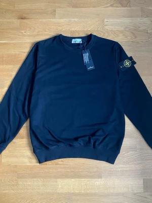Mörkblå stickad tröja Stone Island - Snygg mörkblå stickad tröja från Stone Island med klassisk patch på vänster ärm. Tröjan har rund halsringning, ribbade muddar och en stilren, enkel design. Perfekt för dig som gillar streetwear och vill ha något tidlöst i garderoben.
