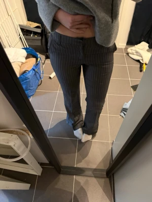 Grå kostymbyxor med vita ränder - Säljer ett par grå kostymbyxor med smala vita kritstrecksränder. Byxorna har rak passform, normal midja och klassisk knappgylf. Perfekta för en stilren och trendig look.