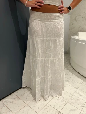 Vit långkjol med resårmidja - Säljer en vit långkjol med bred resår i midjan. Kjolen är luftig och har flera sömmar som ger en snygg, bohemisk känsla. Perfekt för varma dagar och enkel att styla med crop tops eller linnen.