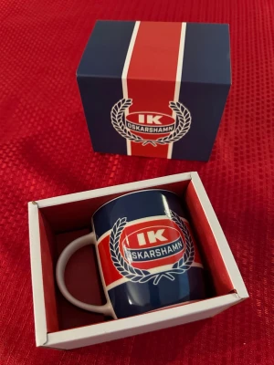 IK Oskarshamn mugg med presentask - Snygg mugg i keramik med IK Oskarshamn-logga i rött, blått och vitt. Muggen har tryck även på handtaget med årtalet 1970. Levereras i matchande presentask med samma färger och klubbmärke. Perfekt för hockeyfans och samlare.