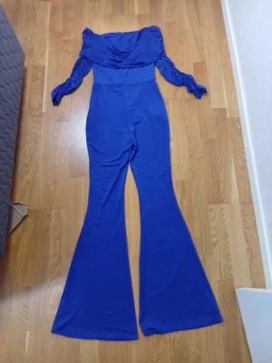 Blå off shoulder jumpsuit - Snygg blå jumpsuit med off shoulder-design och vida ben. Ärmarna är långa och puffiga, vilket ger en trendig look. Materialet ser stretchigt och mjukt ut, perfekt för dig som vill sticka ut. Passar dig som gillar statement-plagg och vill ha något unikt i garderoben.