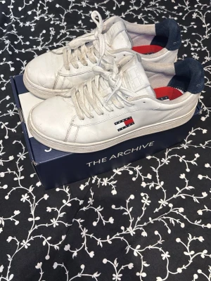 Vita Tommy Hilfiger sneakers - Snygga vita sneakers från Tommy Hilfiger med klassisk logga på sidan och tungan. Skorna har lågt skaft, vita snören och detaljer i marinblått och rött på insidan. Tillverkade i läder för en clean och stilren look.