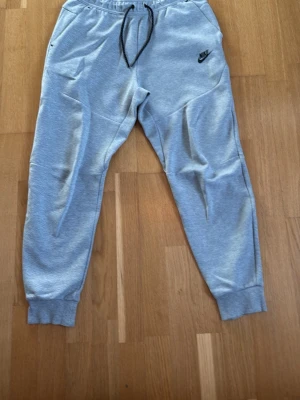Gråa tracksuitmjukisbyxor från Nike - Snygga grå mjukisbyxor från Nike med svart logga på vänster ben. Byxorna har resår i midjan med svart dragsko, två sidofickor och ribbade muddar vid bensluten. Perfekta för chill dagar eller träning. Tillverkade i mjukt och bekvämt material.