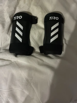 Svarta benskydd Adidas Tiro - Säljer ett par svarta benskydd från Adidas Tiro-serien. De har vita ränder och justerbara kardborreband för bra passform. Perfekta för fotbollsträning eller match. Snygg och sportig design med klassiska Adidas-detaljer.