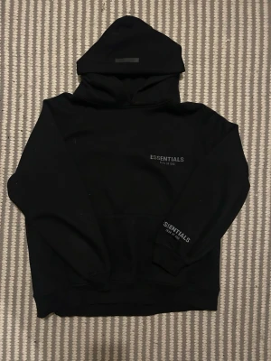 Svart Essentials Fear of God hoodie - Säljer en svart hoodie från Essentials Fear of God med tryck på bröstet och ärmen. Klassisk oversized passform, stor huva och magficka framtill. Tillverkad i mjukt bomullsmaterial som är perfekt för en chill och stilren look.
