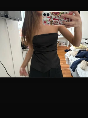 Svart axelbandslös tubtopp - Snygg svart tubtopp med omlott-detalj och asymmetrisk nederkant. Toppen är axelbandslös och har en stilren, elegant look som passar perfekt till både jeans och kjol. Materialet känns mjukt och följsamt mot kroppen.