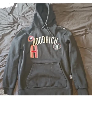 Svart hoodie från Hoodrich med patchar - Svart hoodie från Hoodrich med broderade patchar och tryck på bröstet. Stor ficka framtill och justerbar huva med snören. Mjuk insida och ribbade muddar. Perfekt för en streetwear-look.