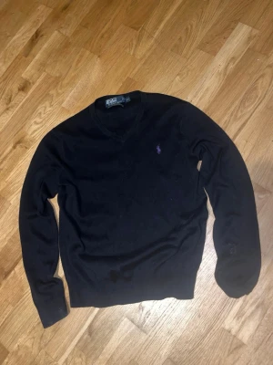 Ralph Lauren - Klassisk svart stickad tröja från Polo Ralph Lauren med lila broderad logga på bröstet. Tröjan har V-ringning och långa ärmar. Perfekt att matcha med jeans eller skjorta för en clean look. Storlek S. Nypris 1499kr