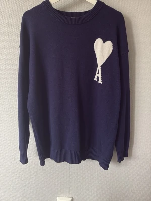 Mörkblå stickad tröja från Ami - Snygg mörkblå stickad tröja från Ami Alexandre Mattiussi med vitt hjärta och bokstaven A på bröstet. Tröjan har rund halsringning, långa ärmar och ribbade muddar. Perfekt för dig som gillar stilrena detaljer och trendig design. Riktigt bra skick knappt använd!