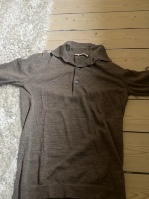 Brun stickad pikétröja A Day's March - Snygg brun stickad pikétröja från A Day's March med klassisk krage och tre knappar framtill. Tröjan är tillverkad i mjukt material och har långa ärmar samt ribbade muddar vid ärmslut och nederkant. Perfekt för en stilren och ”grisch” look