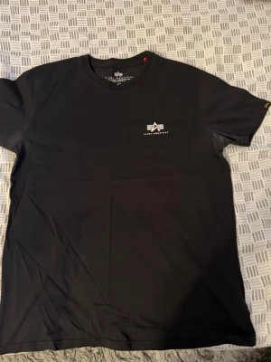 Svart t-shirt från Alpha Industries - Snygg svart t-shirt från Alpha Industries med liten vit logga på bröstet. Klassisk rund hals och korta ärmar. Tillverkad i mjuk bomull som känns skön mot huden. Perfekt till jeans eller shorts för en clean och enkel stil. OBS! Finns små vita fläckar vid armhålorna.
