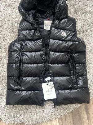 Svart dunväst från Moncler - Snygg svart dunväst från Moncler med glansig finish och klassisk logga på bröstet. Västen har dragkedja framtill, två fickor med dragkedja och huva. Perfekt för lager på lager och streetstyle. Materialet är dunfyllning och yttertyget är blankt.