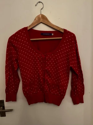 Röd prickig kofta från sunshine - Röd kofta med vita prickar från sunshine. Koftan har rund halsringning, knäppning framtill och ribbade muddar vid ärmslut och nederkant. Perfekt för dig som gillar retrostil och vill ha något färgglatt i garderoben.