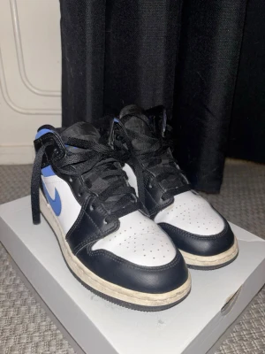 Nike Air Jordan 1 Mid svart/blå/vit - Säljer ett par Nike Air Jordan 1 Mid sneakers i svart, blått och vitt läder. Klassisk siluett med svart snörning, blå Swoosh och Air Jordan-logga på sidan. Perforerad tåbox för extra ventilation och vit mellansula. Perfekta för dig som gillar streetwear.