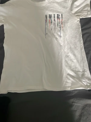 Vit AMIRI t-shirt med färgstänk - Säljer en vit t-shirt från AMIRI med tryck av loggan och färgstänk i blått, svart, rött och grönt både fram och bak. Klassisk passform och rund halsringning. Perfekt för dig som gillar streetwear och vill sticka ut med en unik design.