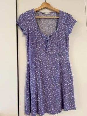 Lila blommig kortklänning från Bershka - Supersöt lila kortklänning från Bershka med små vita blommor, ribbad struktur och knyt i urringningen. Klänningen har korta ärmar och är gjord i mjuk bomull, perfekt för varma dagar.