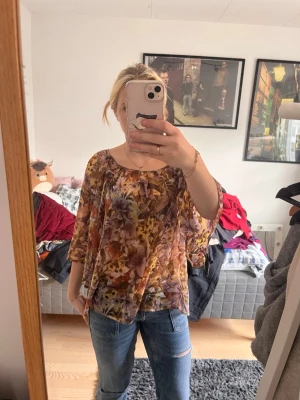 Blommig transparent blus i bruna toner - Säljer en luftig blus med blommigt mönster i bruna, gula och lila nyanser. Blusen har trekvartsärm och rund halsringning. Materialet är tunt och transparent, vilket ger en avslappnad och trendig look. Perfekt att styla med jeans eller kjol.